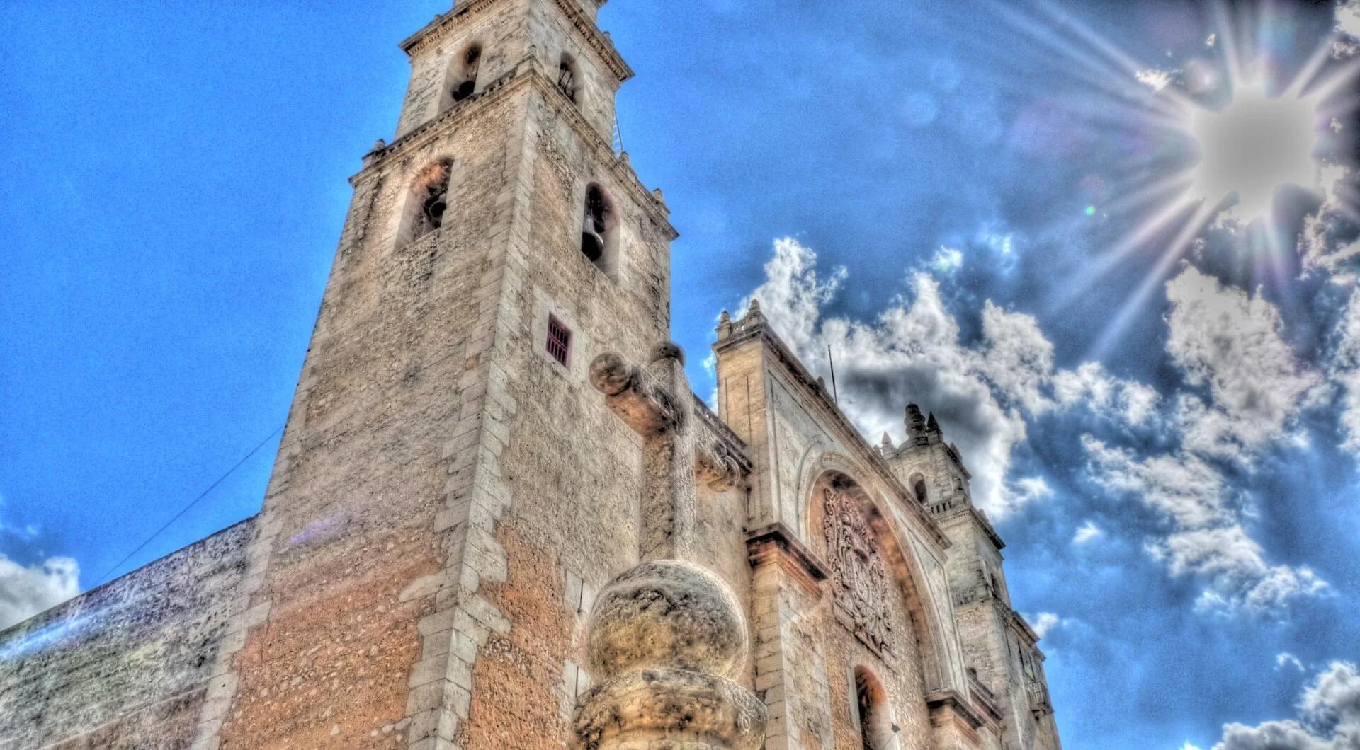 Torre y fachada de piedra de una iglesia colonial de Mérida bajo un cielo azul con sol