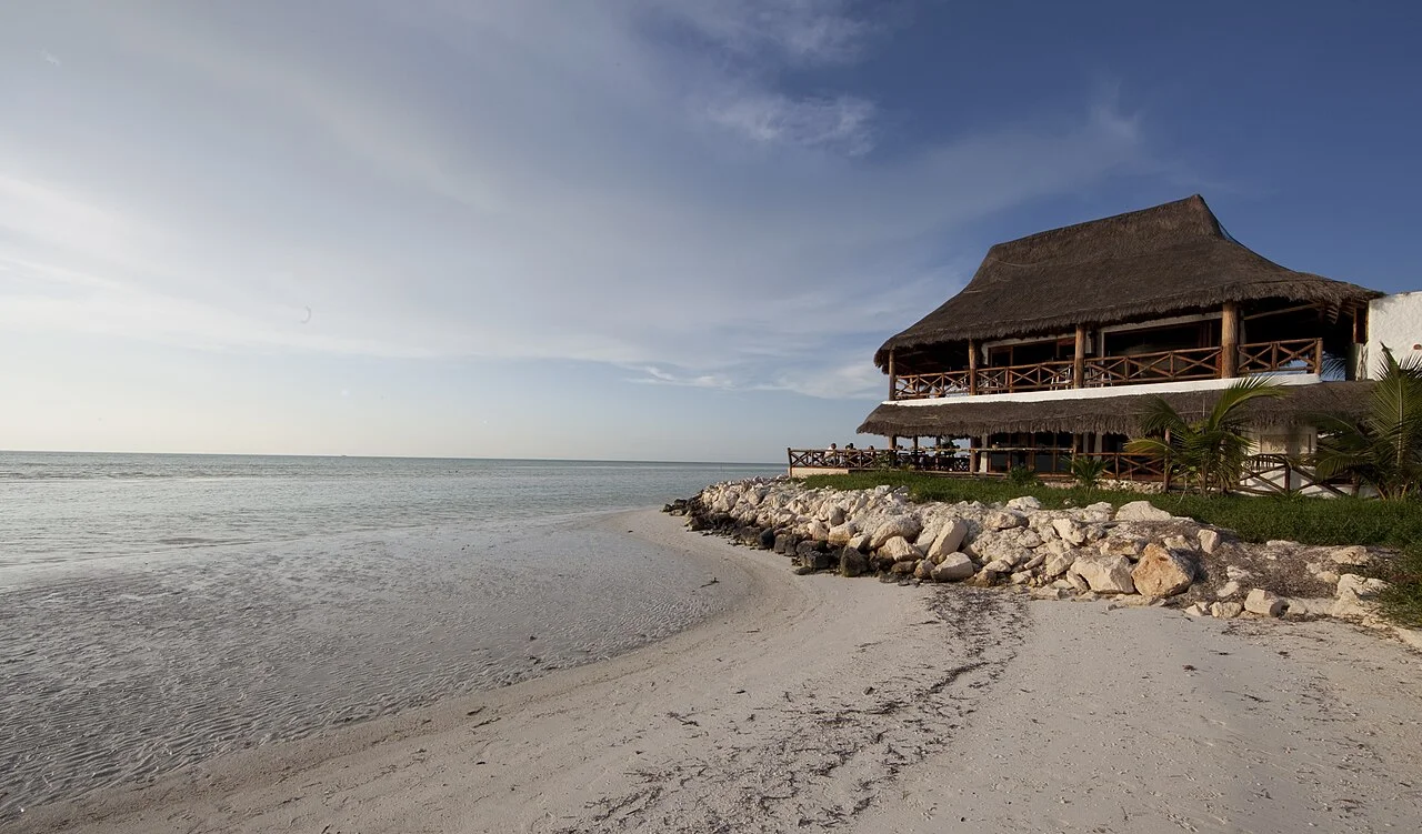 Edificio con techo de palapa y muro de rocas frente a una playa de aguas tranquilas en la zona de Holbox