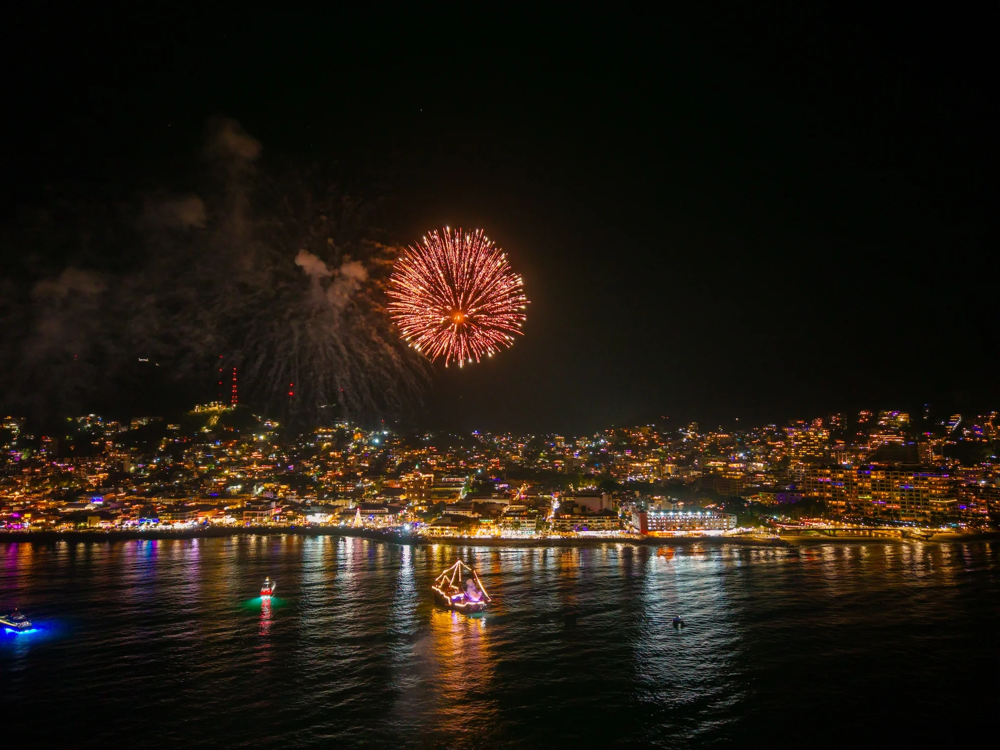 Fuegos artificiales sobre Puerto Vallarta de noche, con la ciudad iluminada y un barco pirata con luces en la bahía
