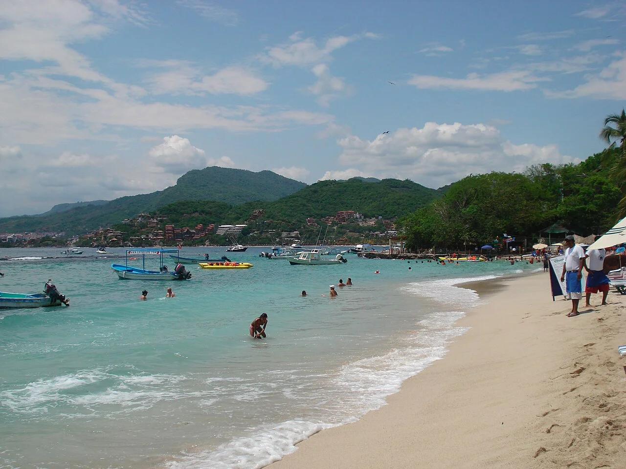 Playa Las Gatas con agua turquesa, lanchas y bañistas, y el pueblo de Zihuatanejo al fondo