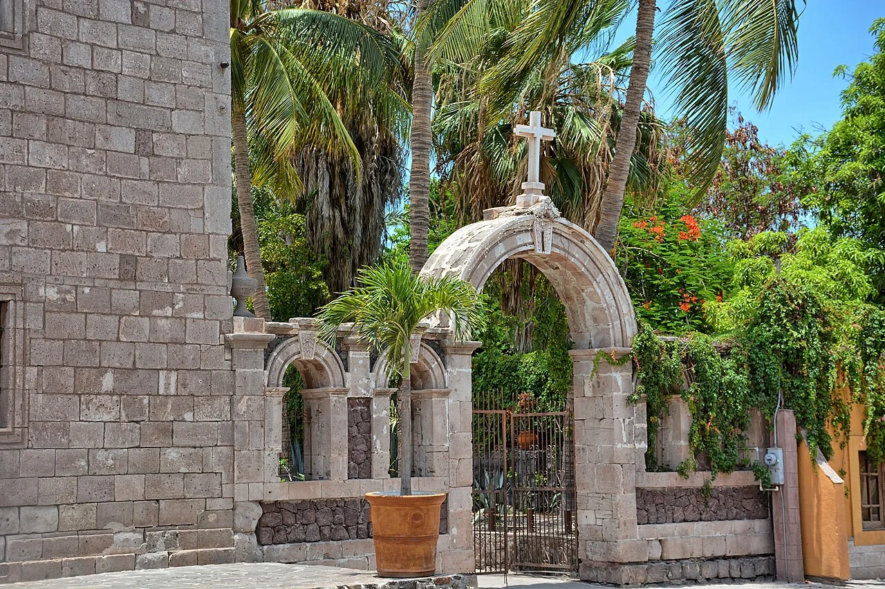 Muro de piedra y arco con cruz entre palmeras en el atrio de la Misión de Loreto, fundada en 1697