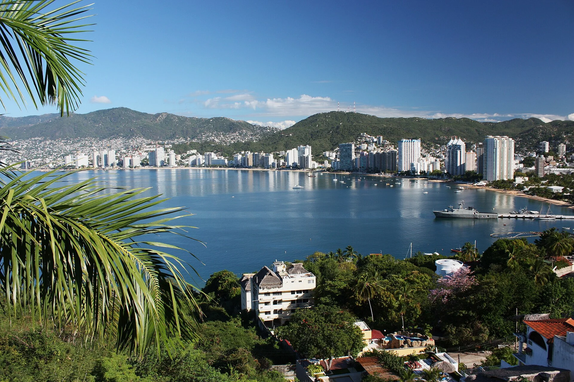 Panorámica de la bahía de Santa Lucía con edificios frente al mar y montañas verdes, enmarcada por palmeras