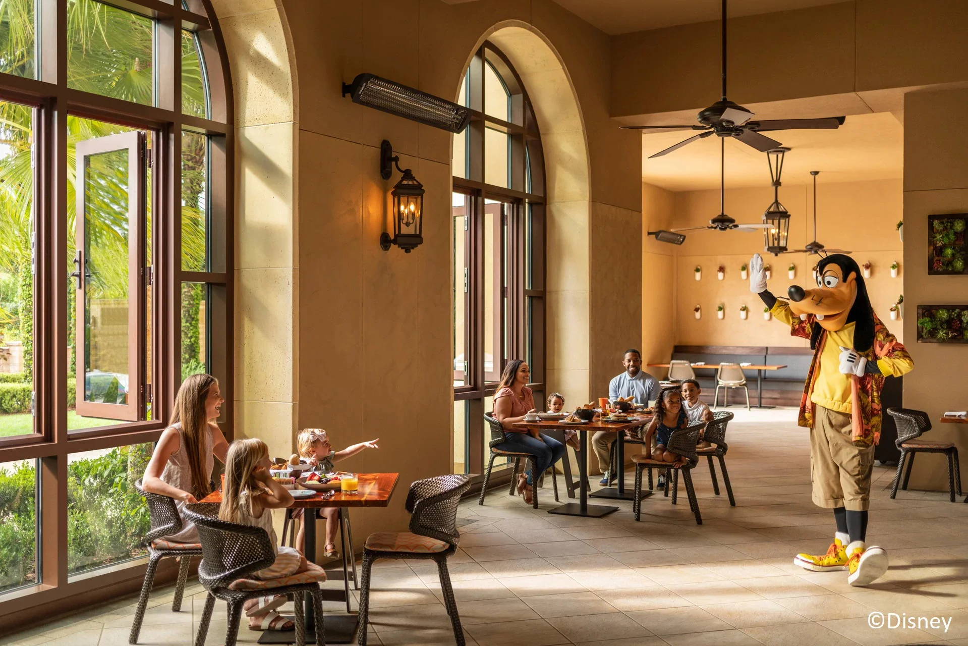Goofy saluda a una familia que desayuna en un comedor con ventanales y palmeras en un hotel de Disney