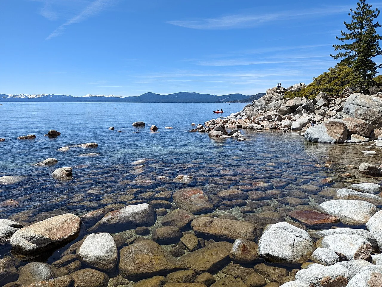 Agua cristalina sobre rocas de granito redondeadas en Sand Harbor, con montañas nevadas al fondo