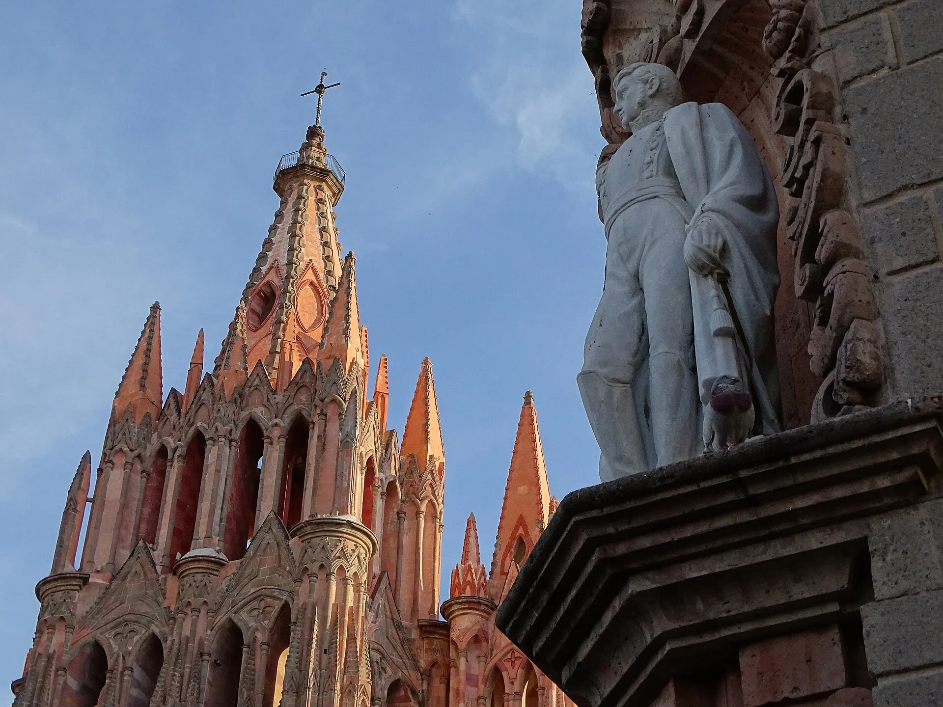 Agujas de la Parroquia de San Miguel Arcángel al atardecer junto a una escultura de piedra en una fachada