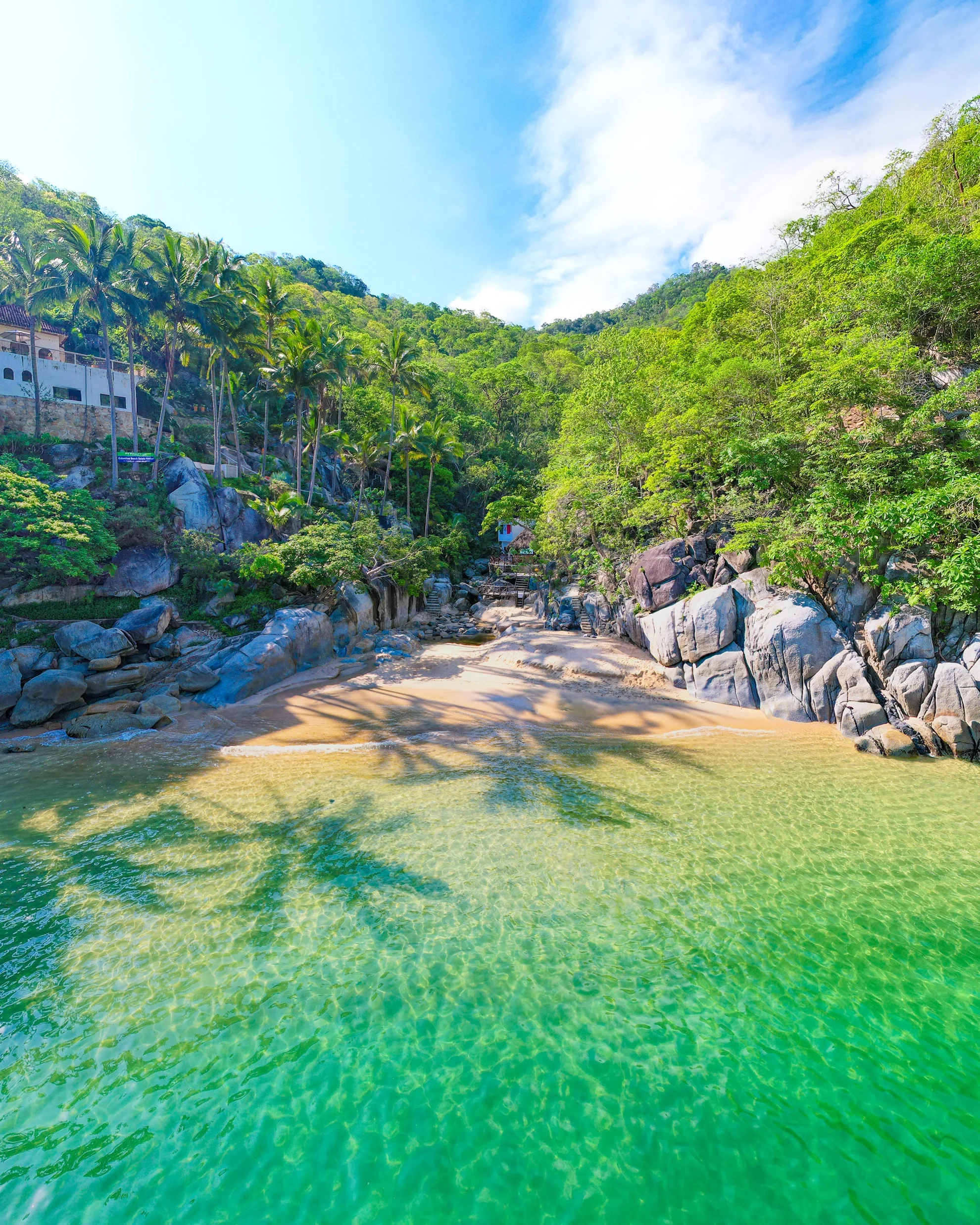 Pequeña caleta de arena con agua verde turquesa, rocas grandes y palmeras sobre la selva de la costa sur de Vallarta