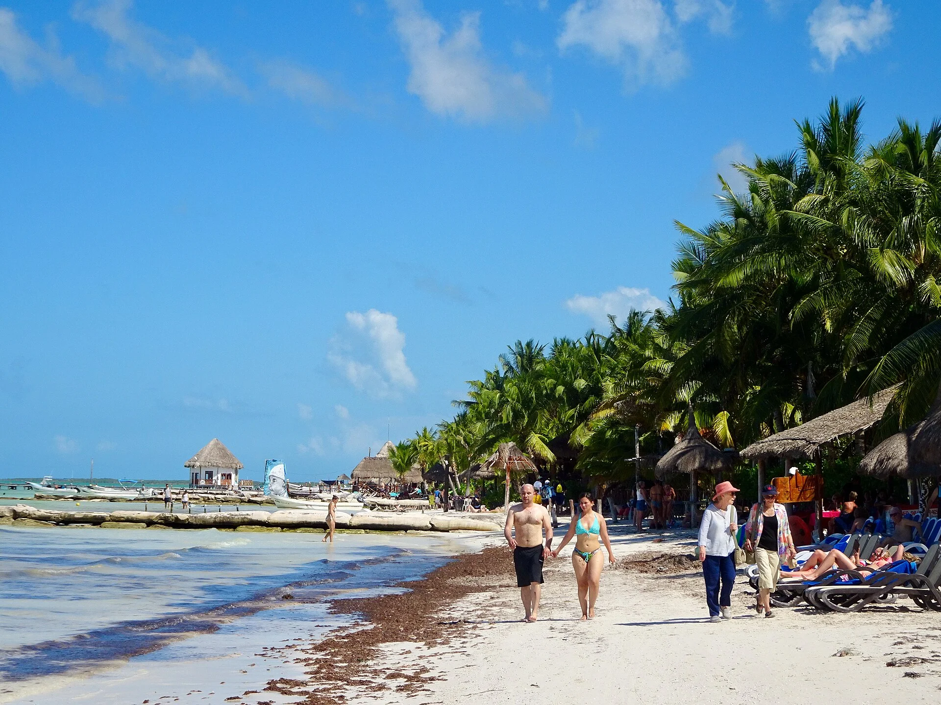 Visitantes caminando por la playa de Holbox junto a palmeras, palapas y camastros