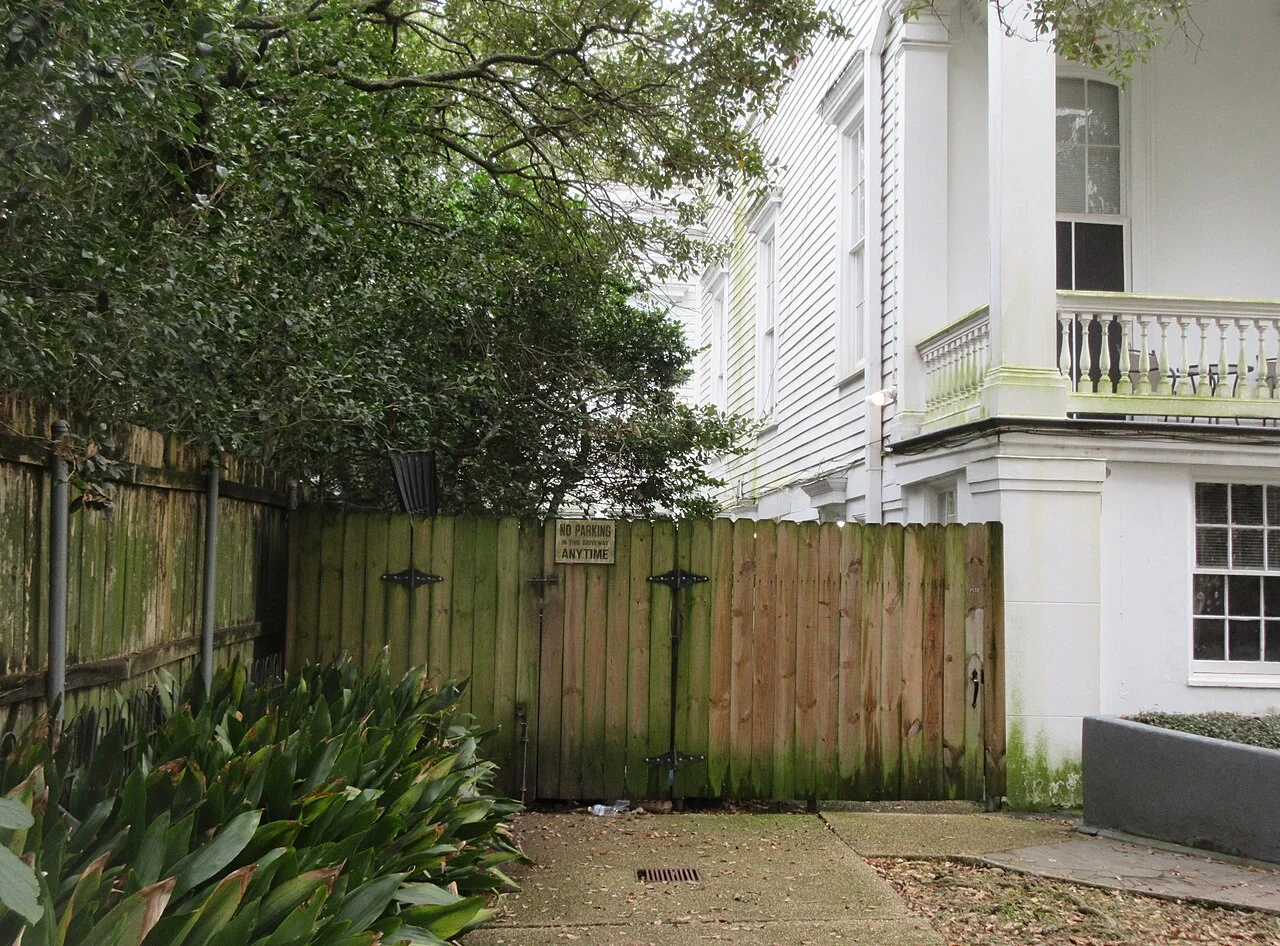 Casona blanca con columnas y barandal junto a una cerca de madera y vegetación en el Garden District