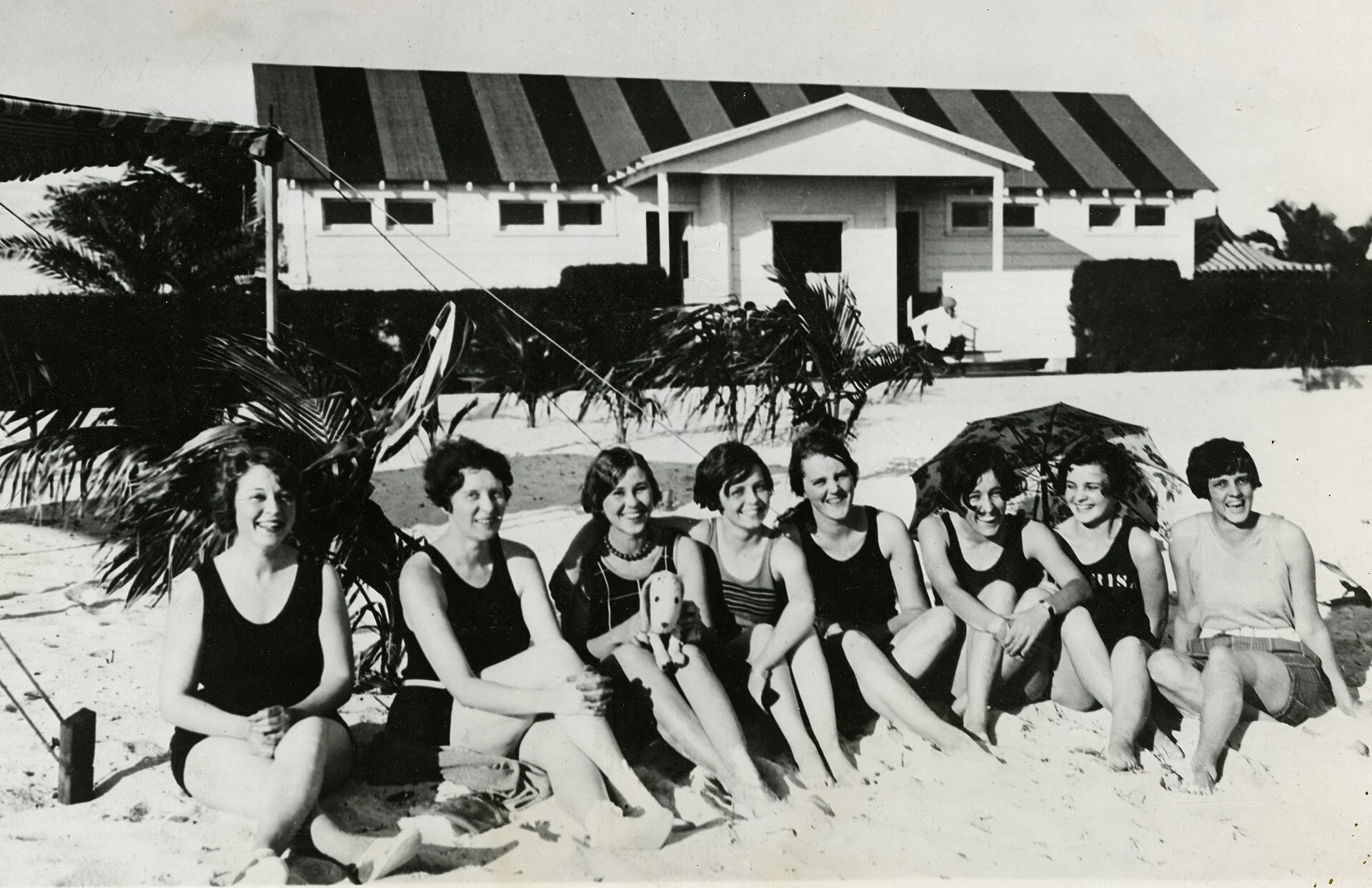 Foto antigua en blanco y negro de mujeres en traje de baño sentadas en la arena frente a un pabellón de playa