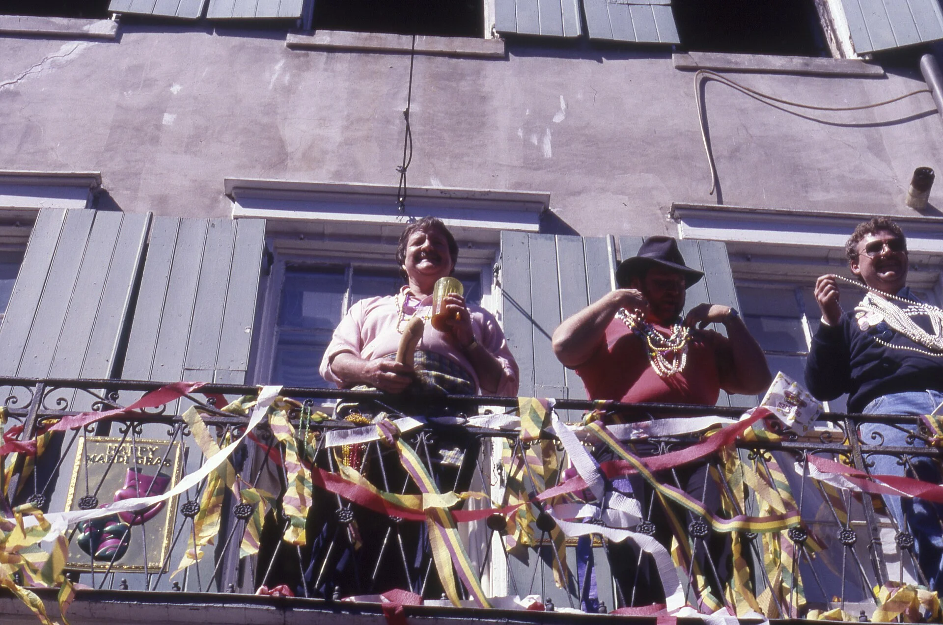 Personas con collares en un balcón adornado con listones dorados, morados y rojos durante Mardi Gras