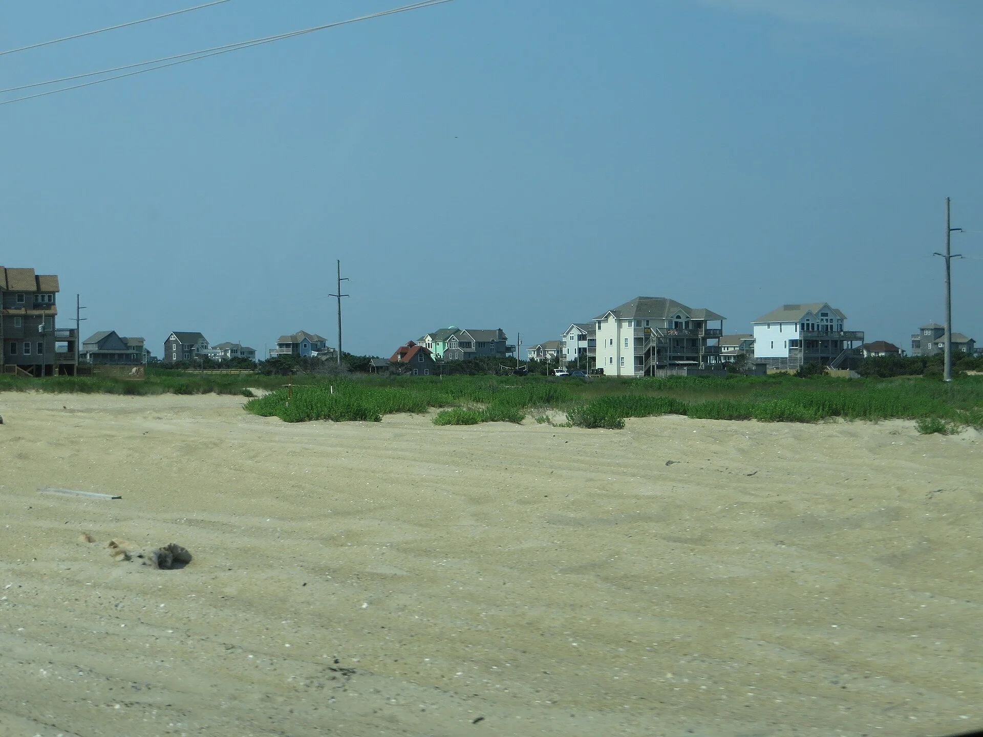Terreno arenoso con pastos costeros frente a una hilera de casas de playa de varios pisos