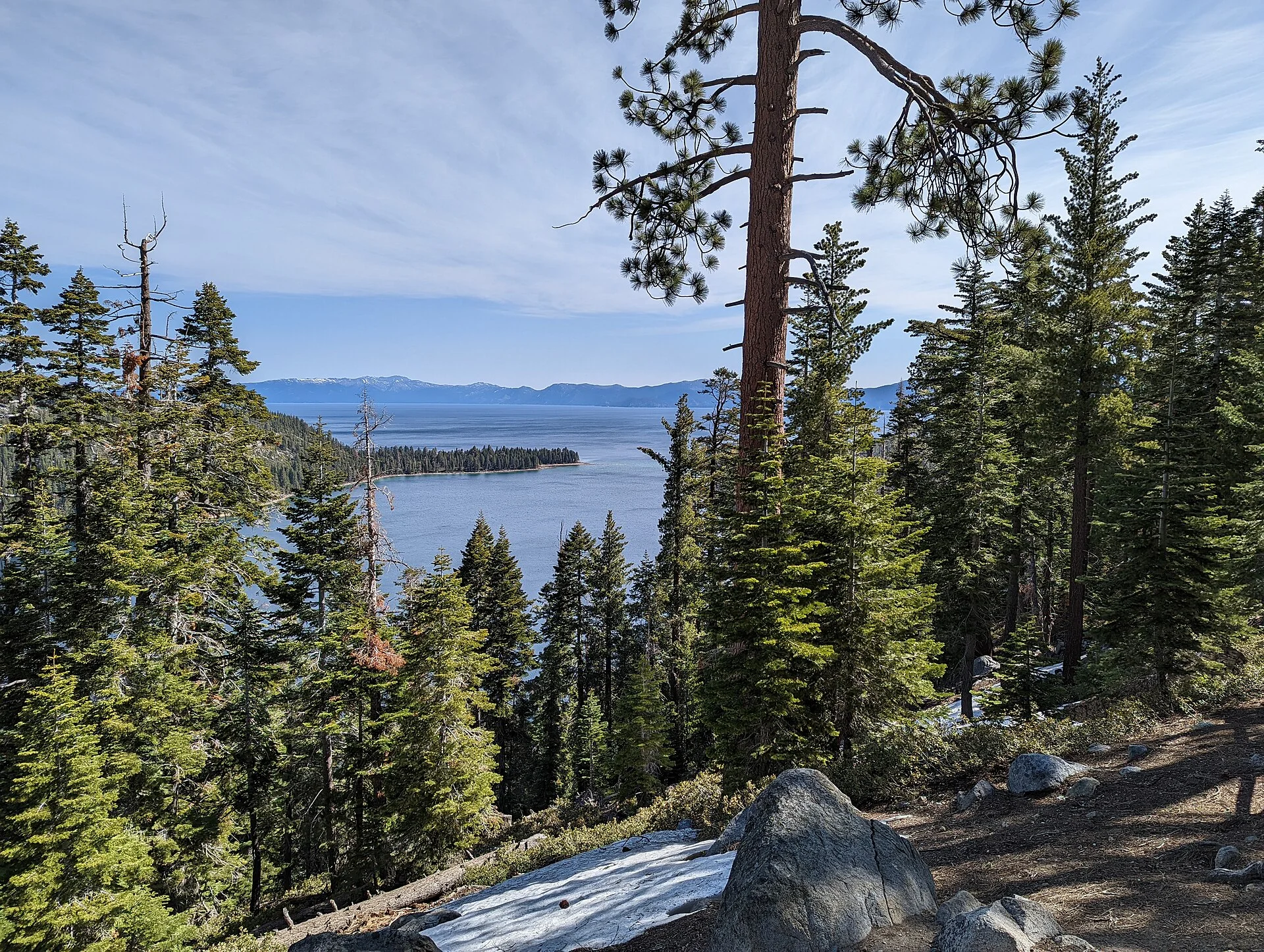 Vista del lago Tahoe entre pinos desde un sendero, con rocas y restos de nieve en primer plano