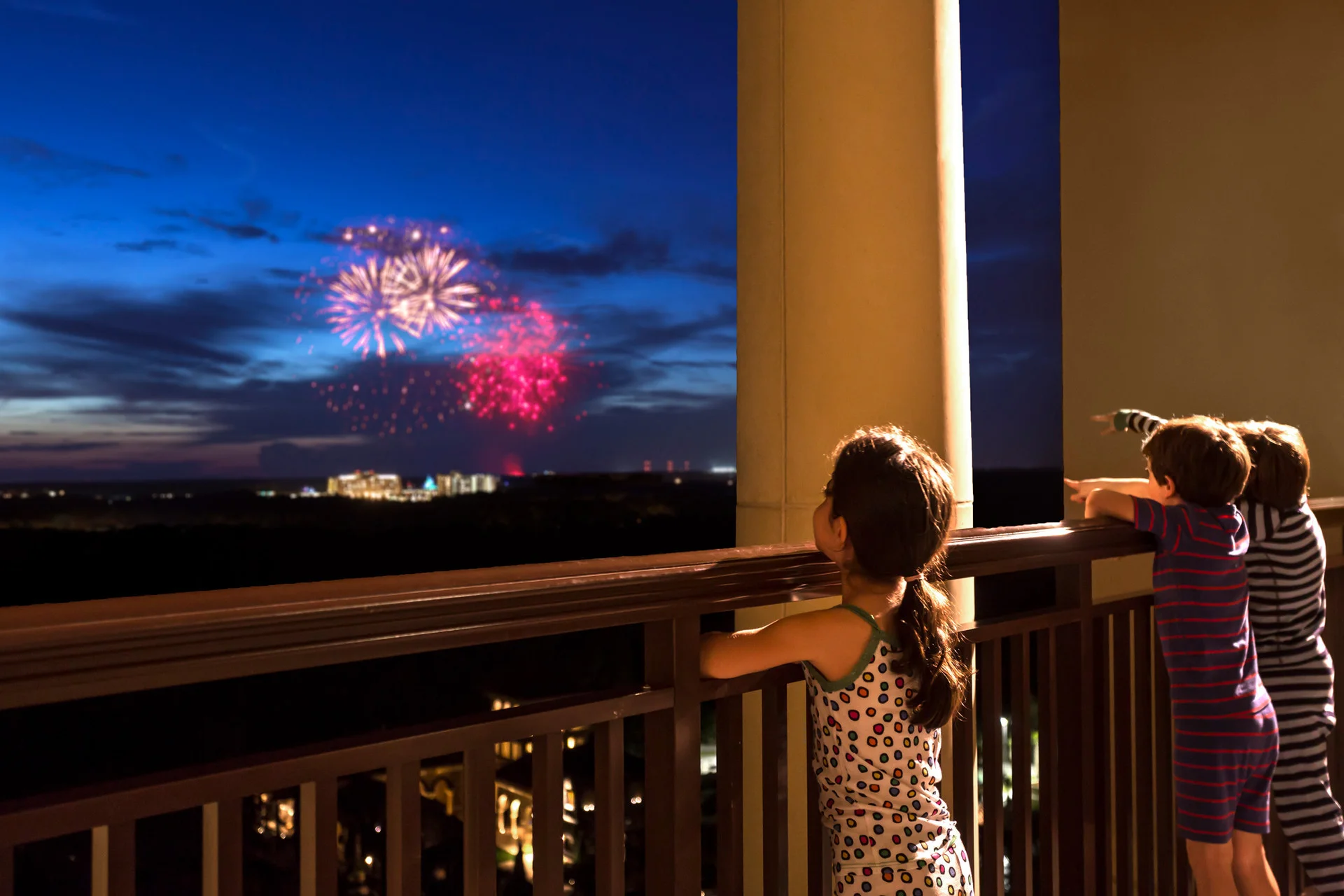 Niños asomados a un balcón de hotel mirando fuegos artificiales de colores sobre el horizonte nocturno de Orlando