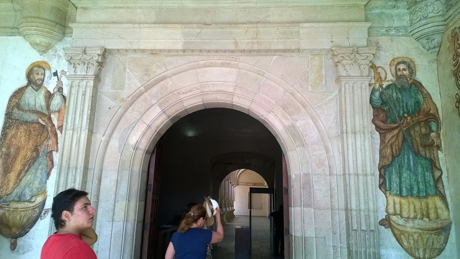 Arco de cantera flanqueado por pinturas murales de santos en un antiguo convento de Oaxaca, con visitantes