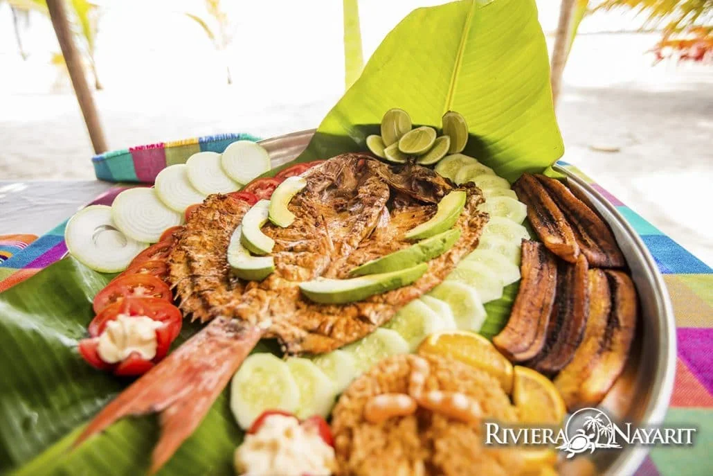 Pescado zarandeado servido sobre hoja de plátano con aguacate, pepino y plátano frito