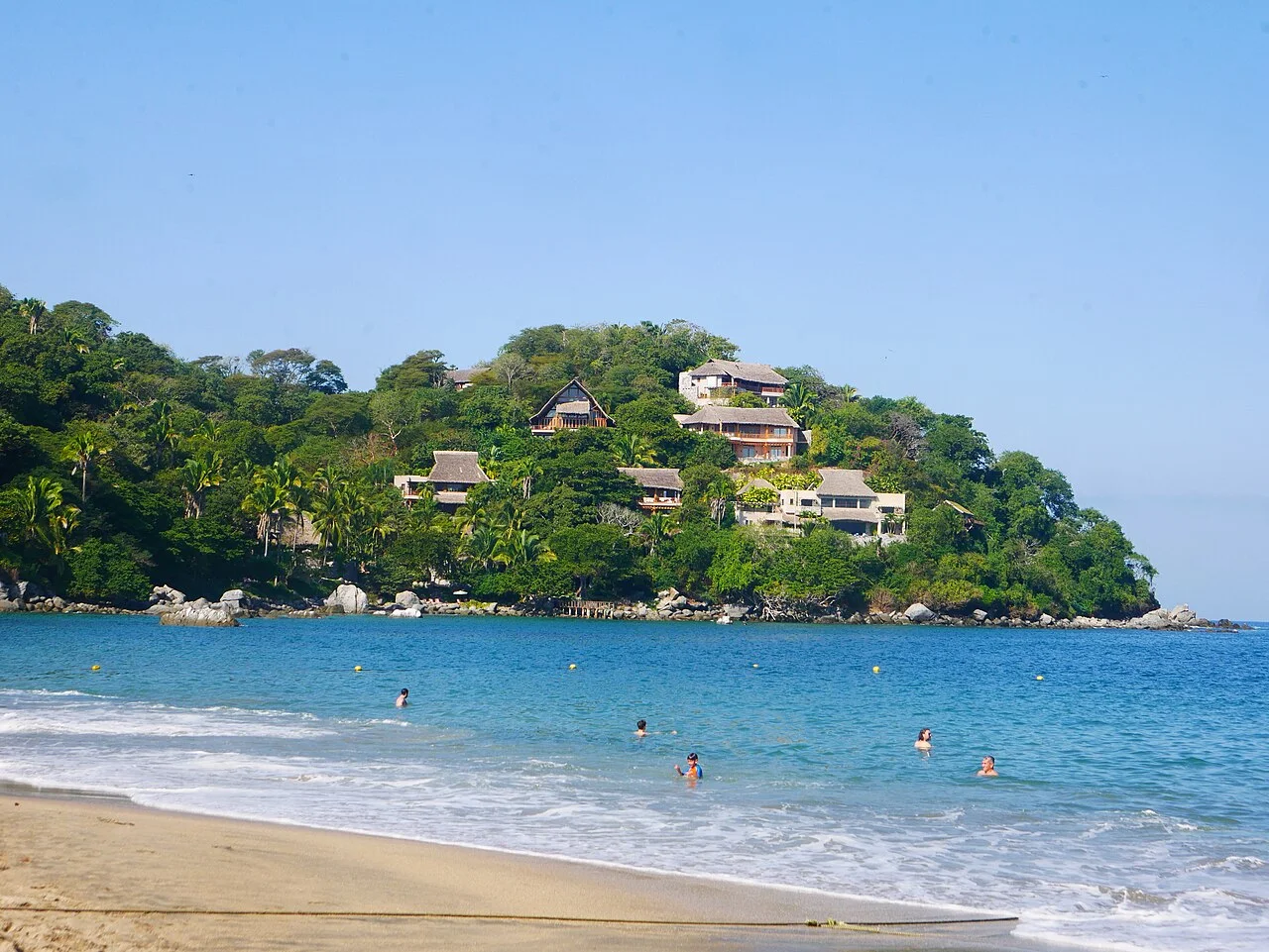 Bañistas en el agua tranquila de Playa de los Muertos con casas entre la selva en la colina