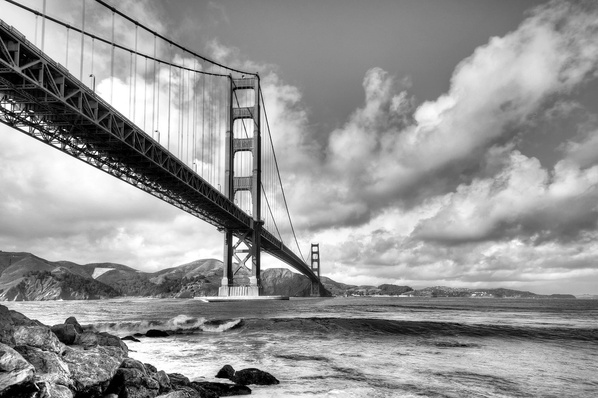 Fotografía en blanco y negro del Golden Gate visto desde abajo, con rocas y nubes dramáticas