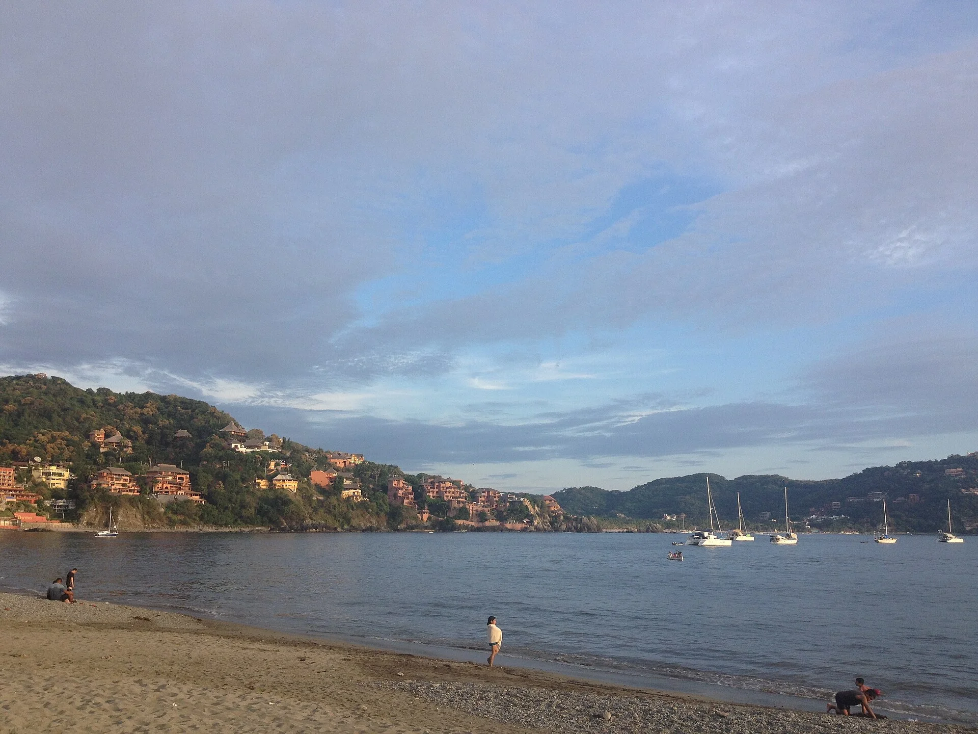 Playa de la bahía de Zihuatanejo al atardecer, con veleros fondeados y casas sobre el cerro