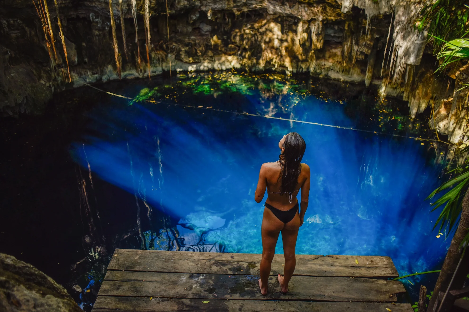 Mujer de pie en una plataforma de madera frente a un cenote iluminado por rayos de luz azul