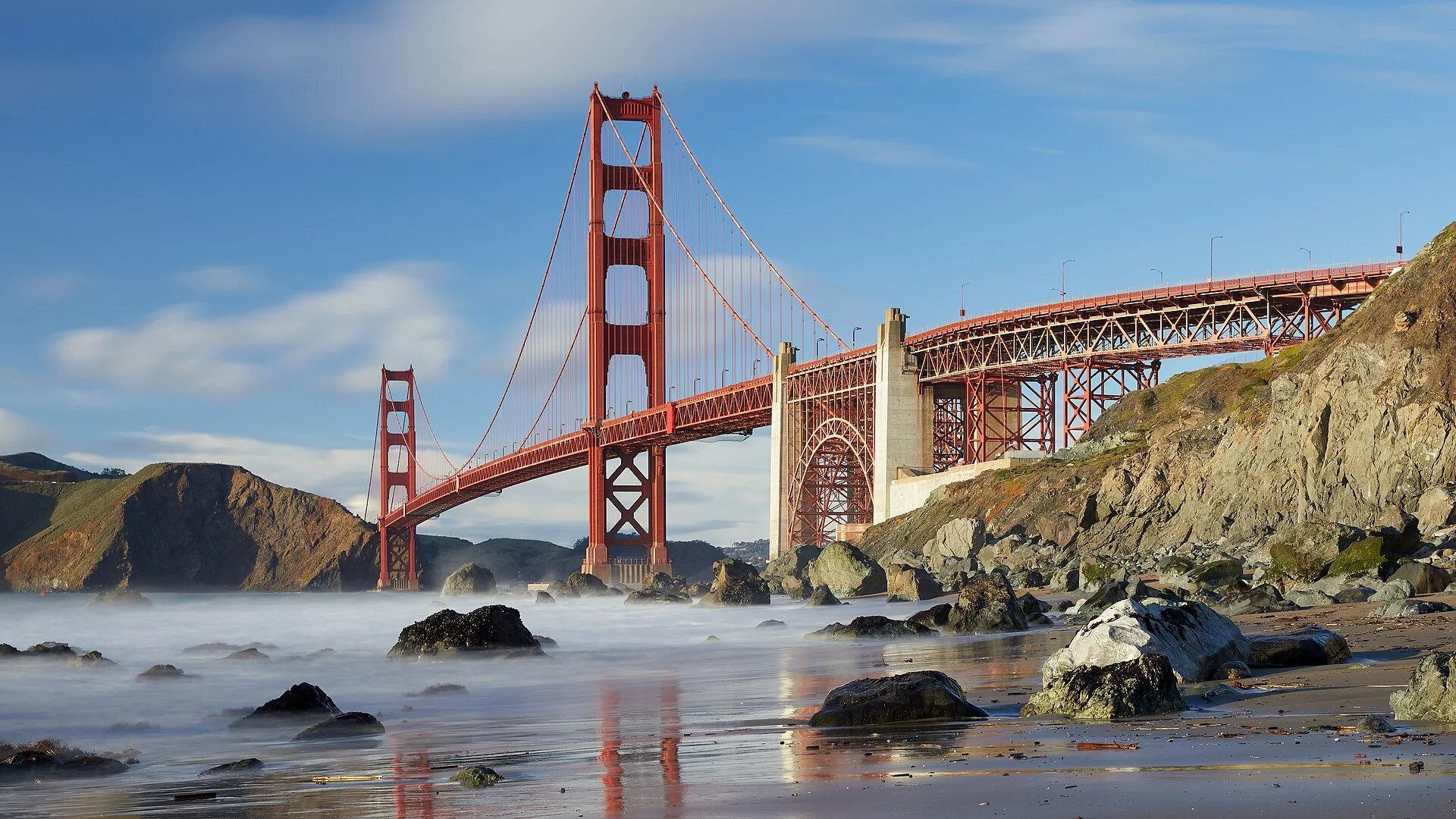 Puente Golden Gate al atardecer desde una playa rocosa, con el mar difuminado por larga exposición