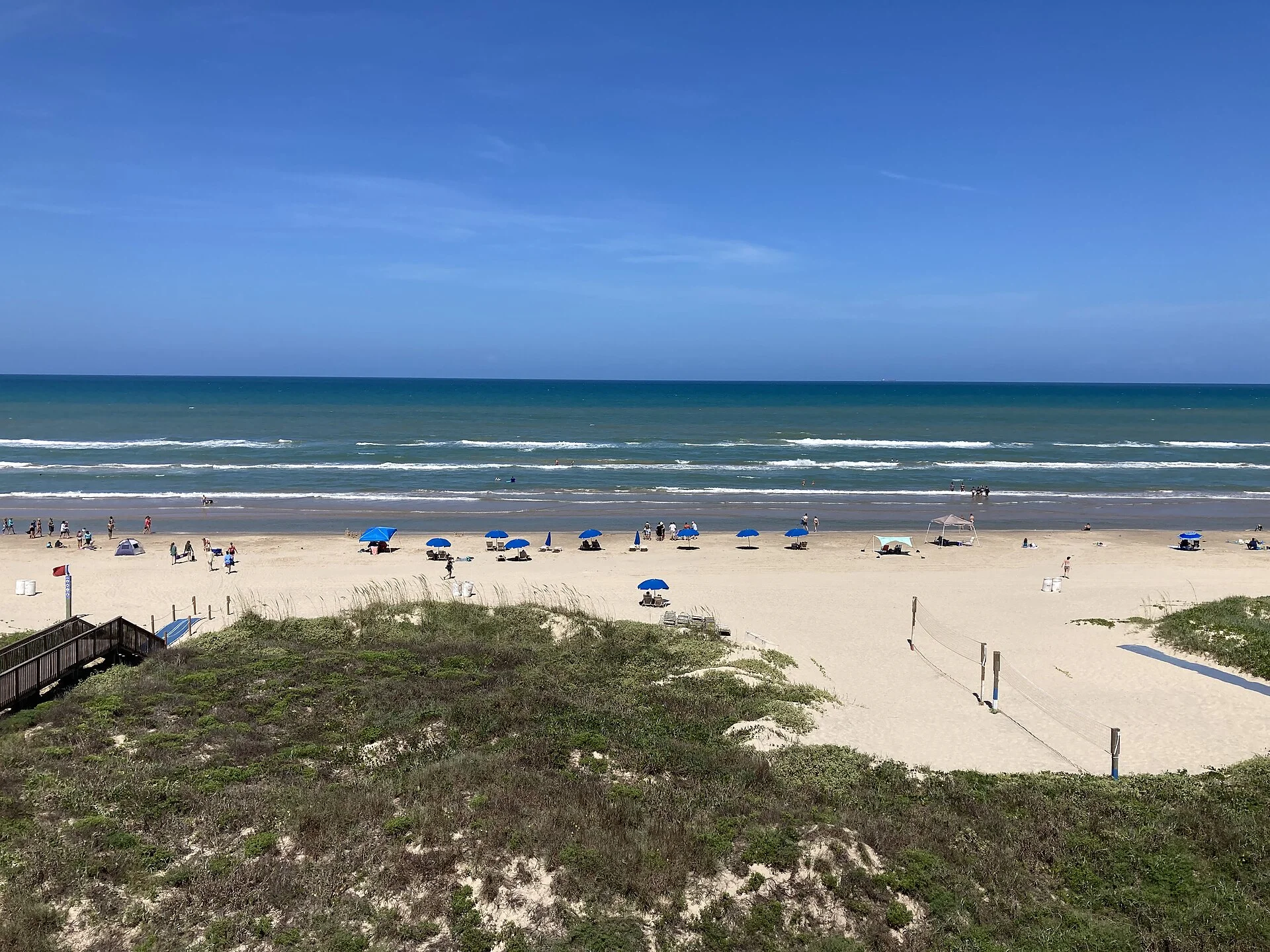 Playa de arena clara con sombrillas azules, dunas con vegetación y mar turquesa en un día despejado