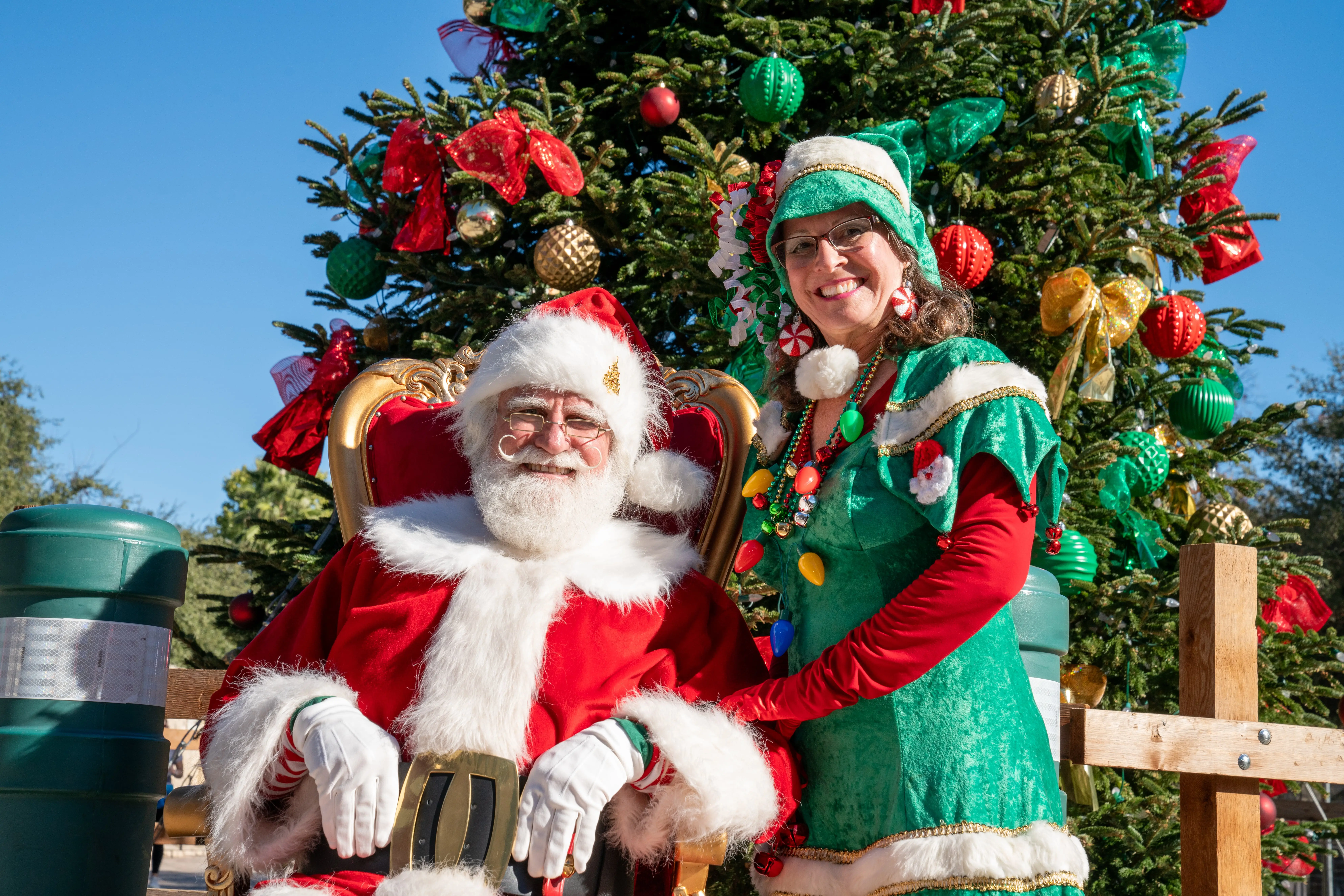 Santa Claus y una elfa sonriendo frente a un gran árbol de Navidad decorado al aire libre